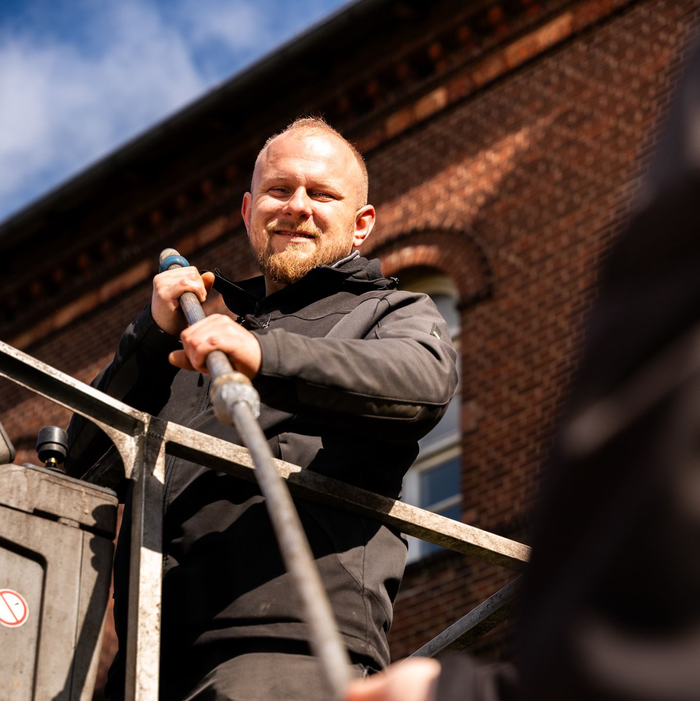 Ein Mitarbeiter steht im Hebekorb und bekommt eine Reinigungsstange von einem Kollegen angereicht